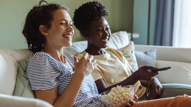 Deux femmes sont confortablement assises sur un canapé avec un bol de pop-corn et regardent la télévision
