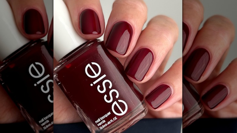 Gros plan d'une personne avec la teinte bordeaux d'Essie peinte sur ses ongles, tenant la bouteille de vernis à ongles