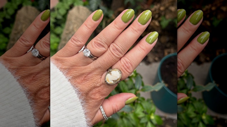 Main ouverte avec divers anneaux montrant du vernis à ongles olive d'Olive & June avec des plantes en pot en arrière-plan