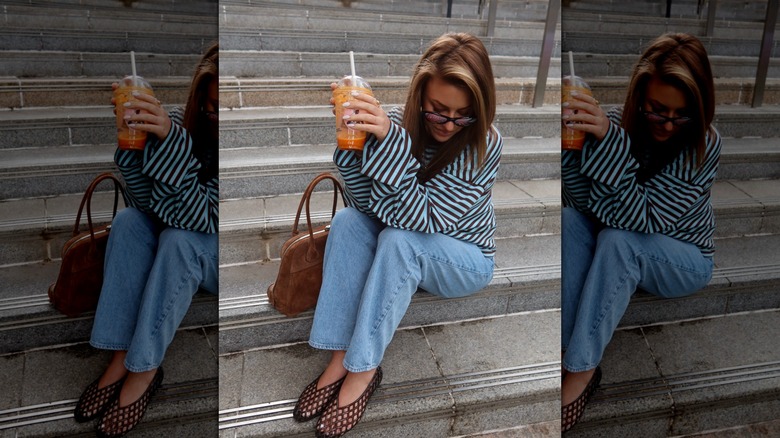 Image d'un influenceur de style tenant un verre et posant pour une photo dans un t-shirt à manches longues à micro-rayures, un jean légèrement délavé et des chaussures en gelée