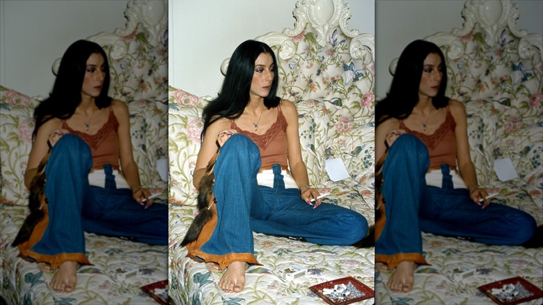 Cher est assise dans son lit, vêtue d'un jean évasé et d'un gilet marron.